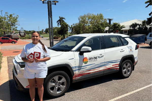 A young woman stands in front of a small white car. She is holding up 2 red P plates