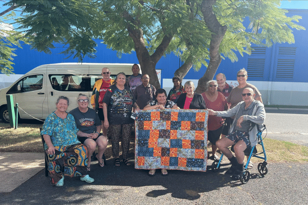A group of people pose in front of a tree. They're holding up a quilt made up of colourful squares