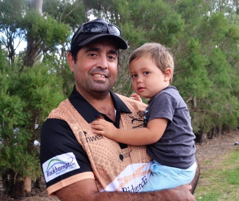 A man wearing a beige cricket shirt holds a young child