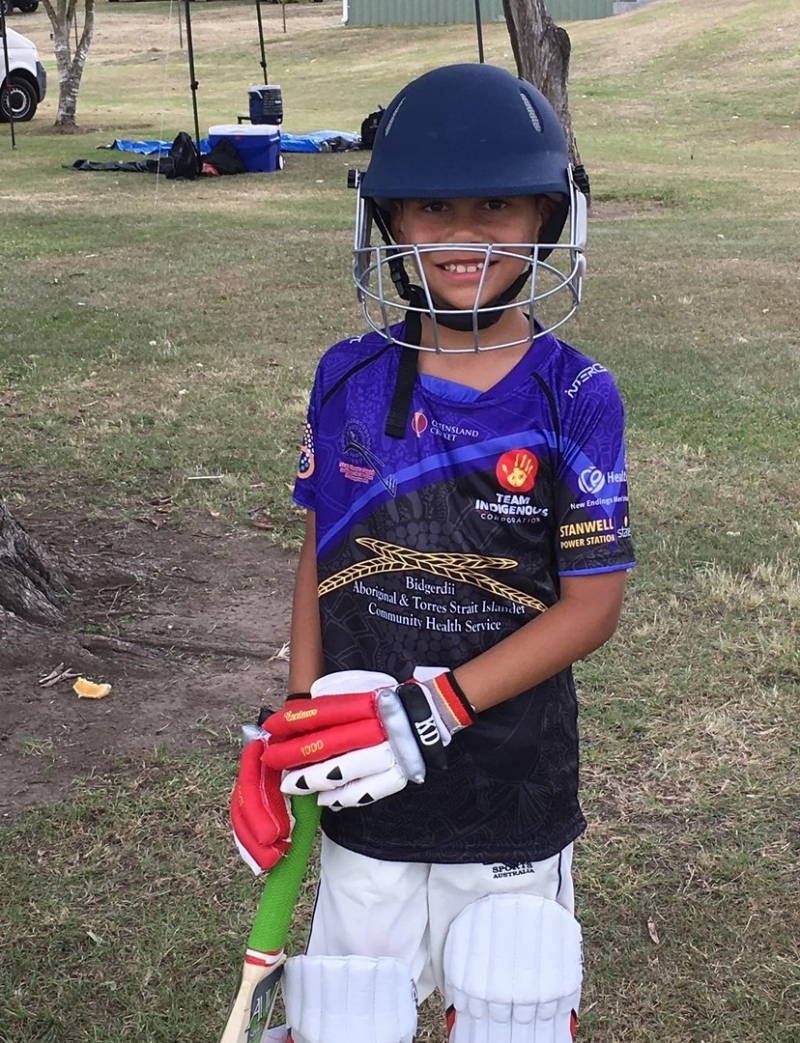 A boy wearing a cricket helmet, gloves and pads, and holding a bat, smiles at the camera