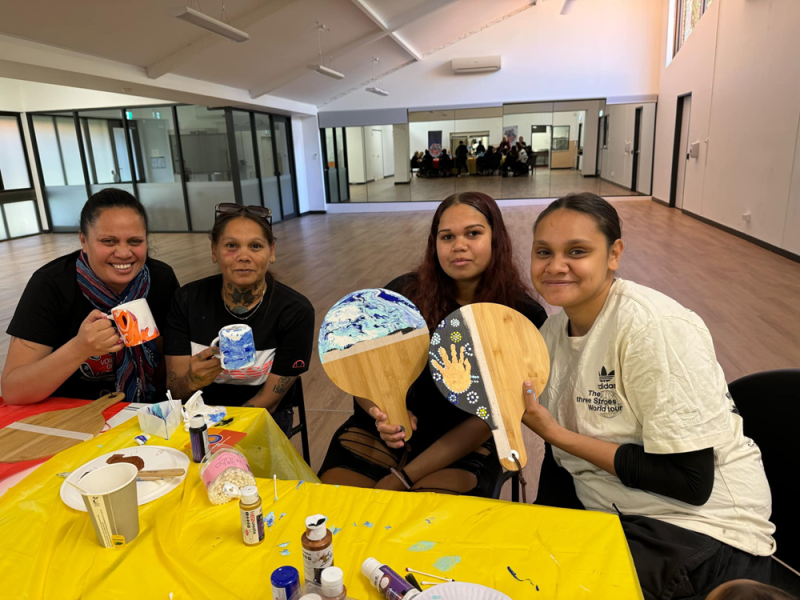 Four women sit in a row at a table that has art supplies on it. The two women on the right are holding up decorated wooden boards. The two women on the left are holding up painted mugs