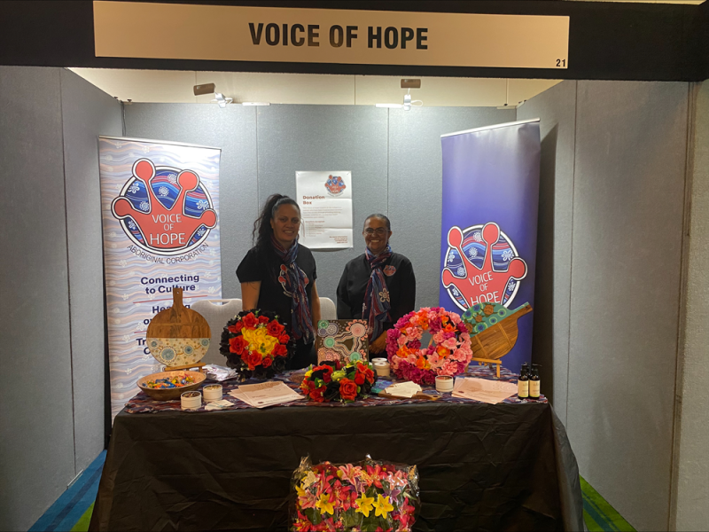 Two women stand behind a table at a stall with Voice of Hope banners on either side and above. On the table are floral wreaths and Aboriginal artwork.