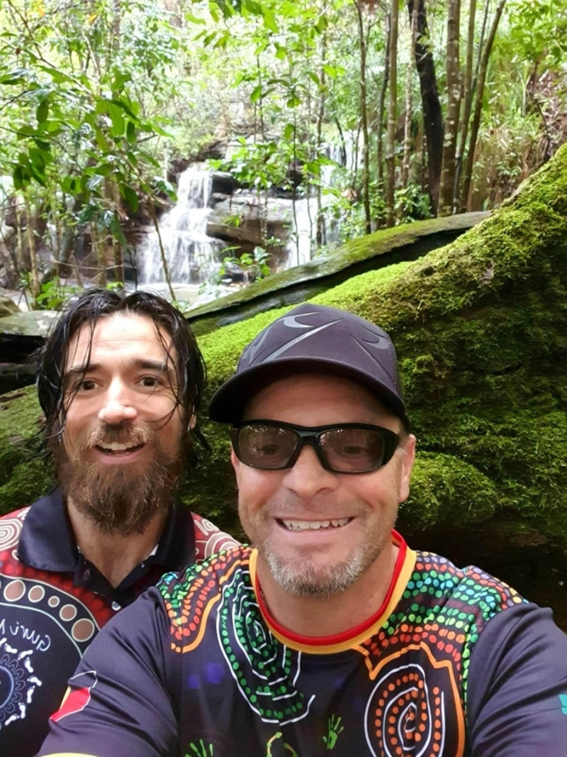 Two men smile at the camera. Behind them are trees and a waterfall