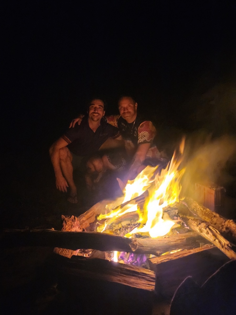 A bonfire in the foreground and 2 men sit closely together in the background