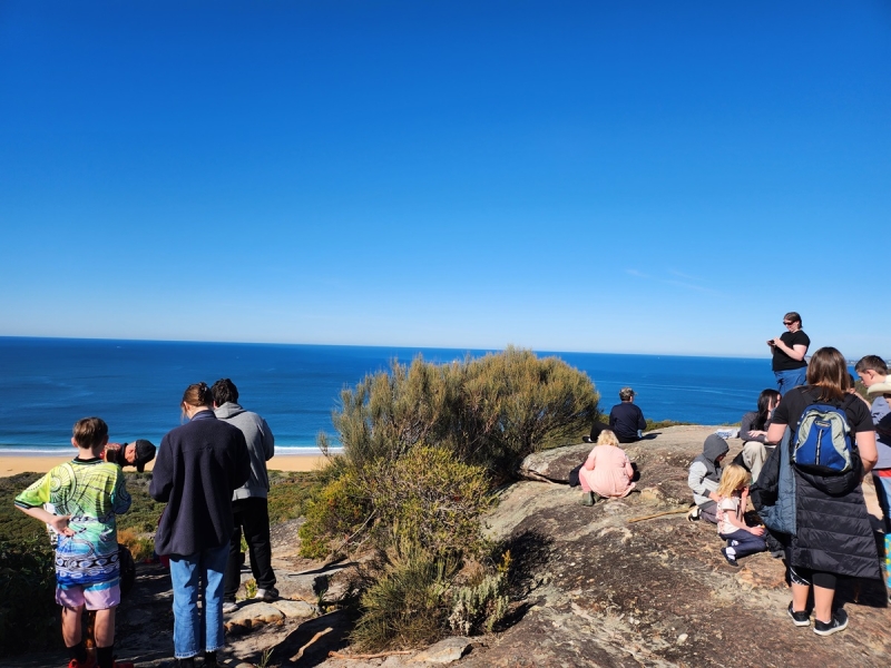 A group of people walk over rocks. In the background is the ocean and blue sky