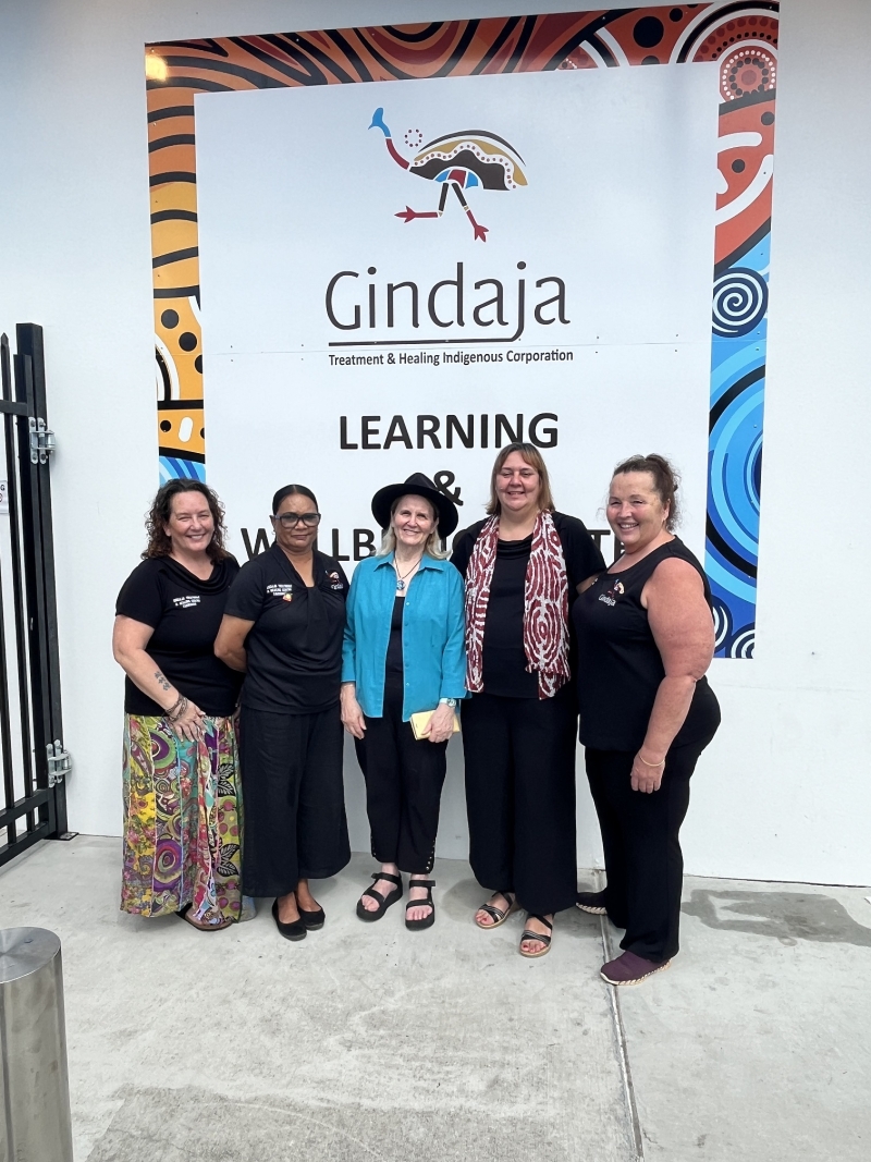 A group of  women stand smiling at the camera. On the wall behind them a large sign says Gindaja