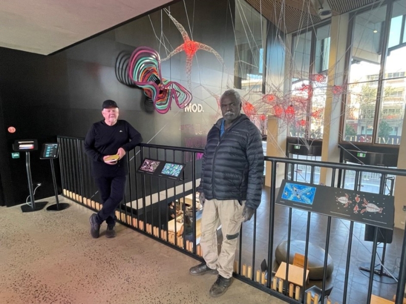 Two men stand on an indoor balcony with an artwork hanging in the space behind them