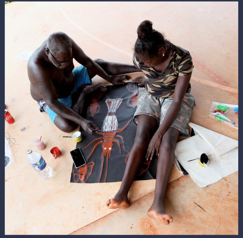 Two people sit on the floor. Between them is a large black piece of paper and the man is painting a crayfish on it