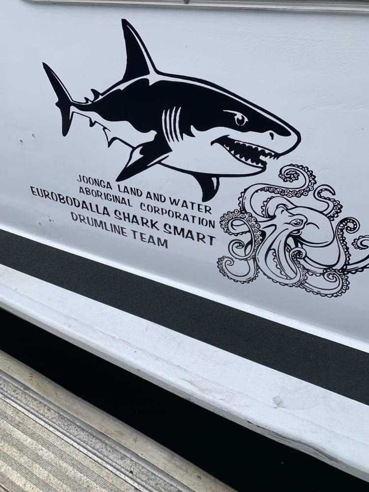 The side of boat with an illustration of a shark and an octopus. The text next to them says Joonga Land and Water Aboriginal Corporation: Eurobodalla Sharksmart Drumline Team