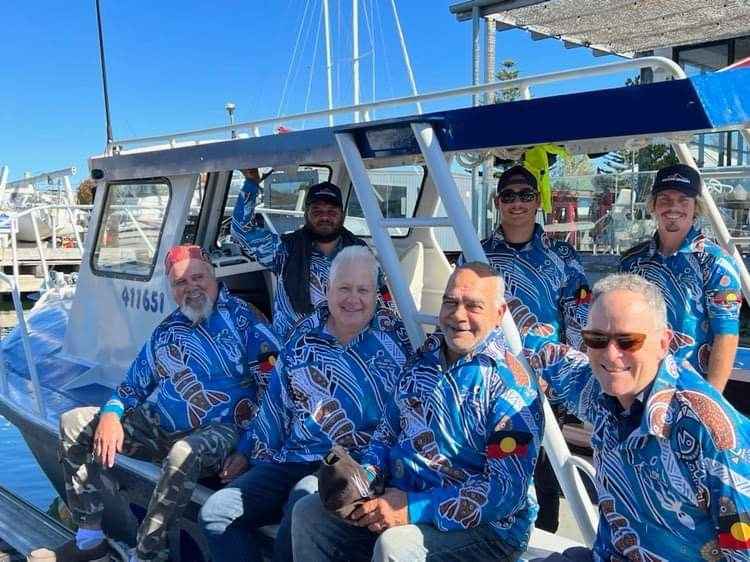 A group of men, all wearing the same blue shirt, pose on the side of a boat and smile at the camera