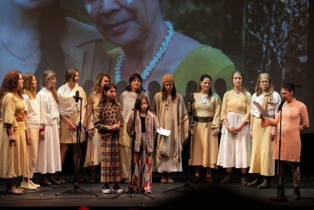 A large group of women stand on a stage. In front of them stand 2 children