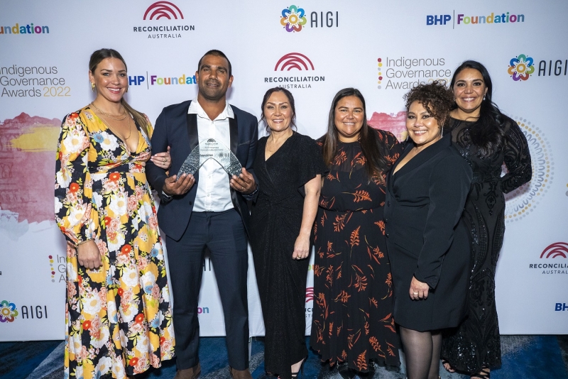 A group of people stands in front of a banner with the Reconciliation Australia logo on it. One person is holding a trophy.