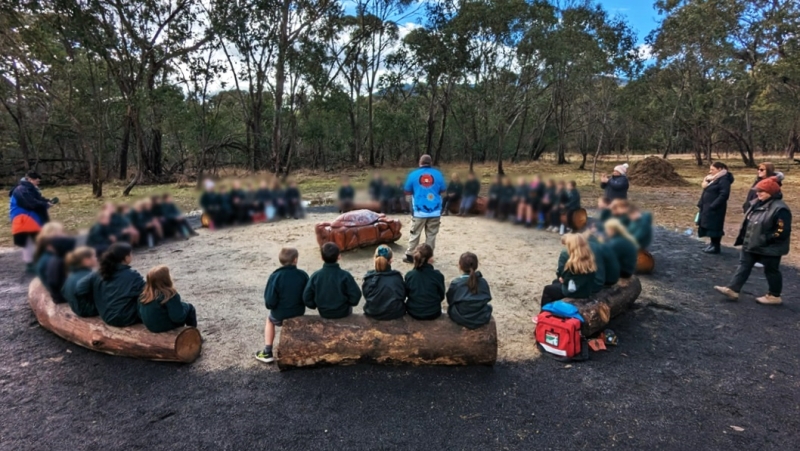 A group of children sit on logs arranged in a large circle. A man in blue stands in the middle of the circle.