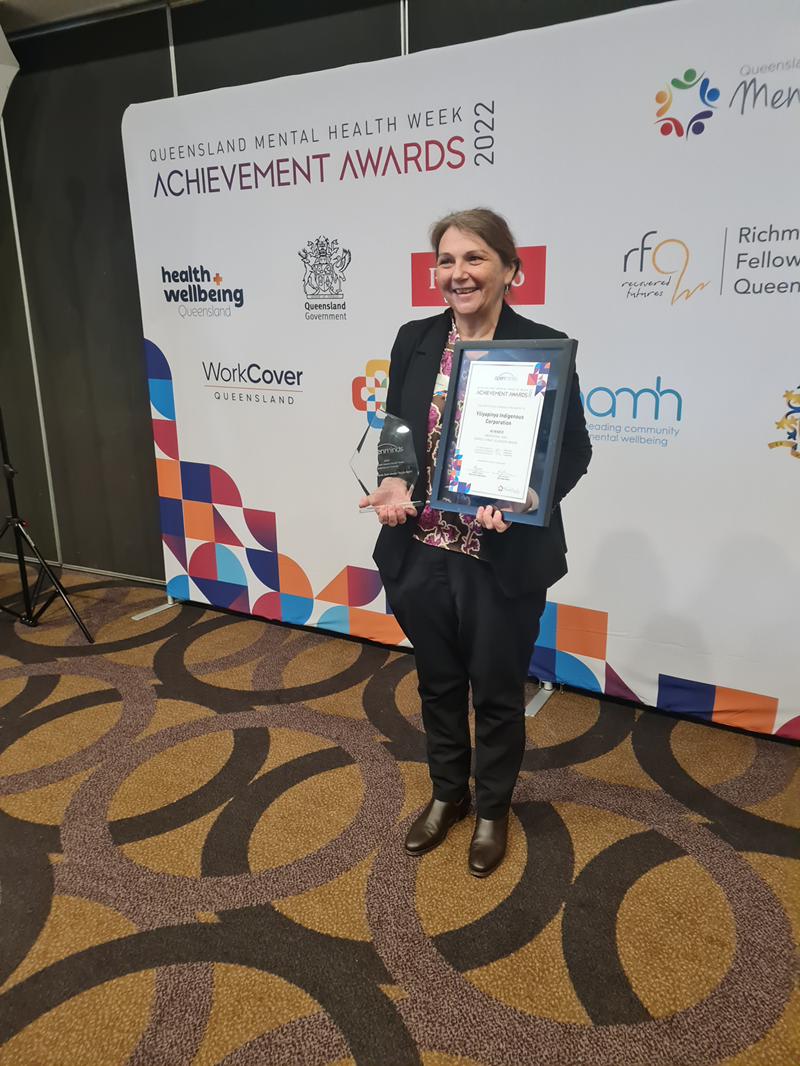 A woman holds a trophy and a certificate. On a banner behind her are the words Queensland Mental Health Week Achievement Awards 2022