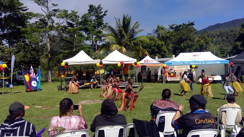 People sit on plastic chairs in the foreground, watching a group of Aboriginal dancers on the grass in front of them. Behind the dancers are temporary marquees