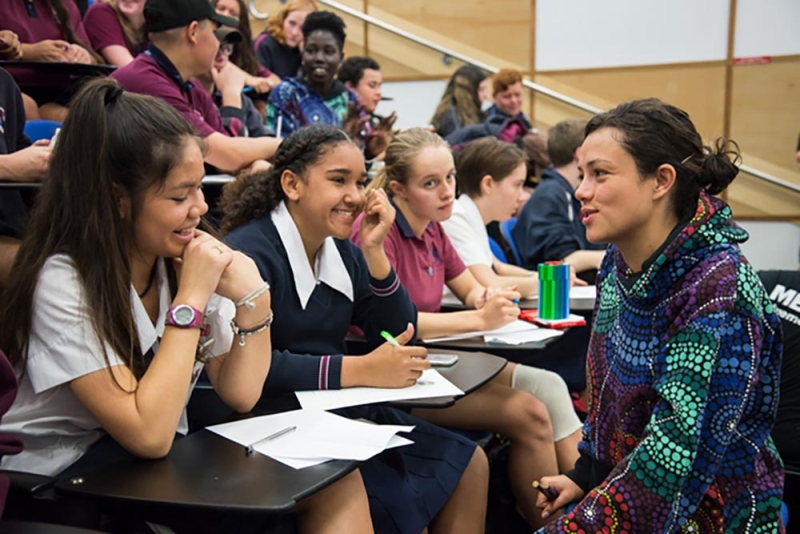 students sit at desks and an adult stands close talking to one of the students