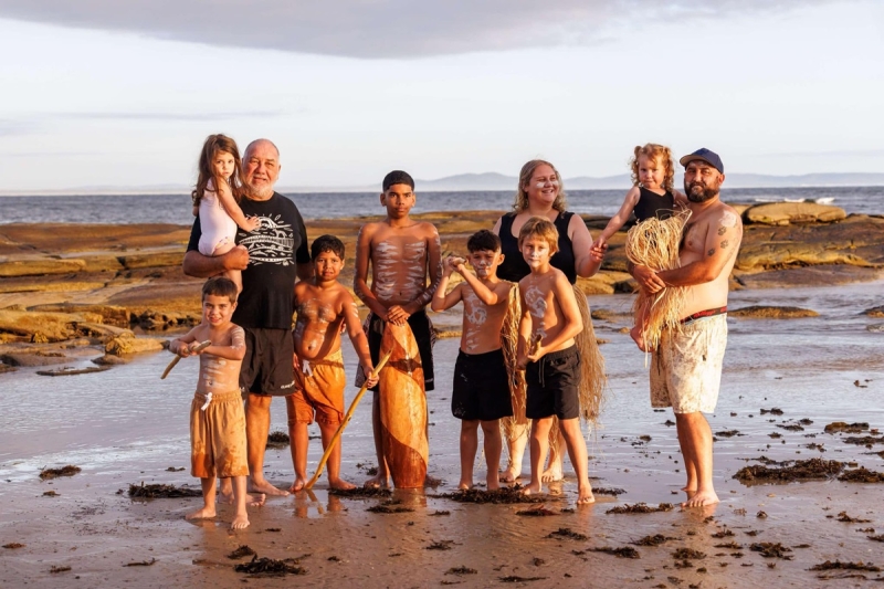 A group of 4 adults and 7 children stands on a beach