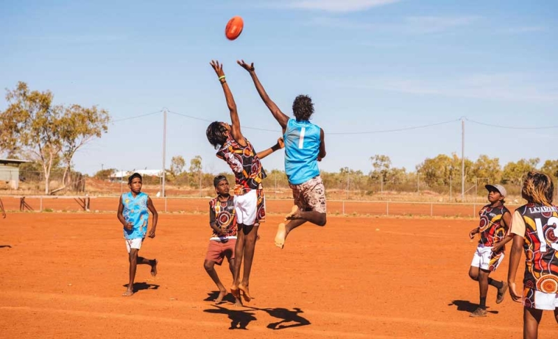 Players jump for a football on a red dirt field