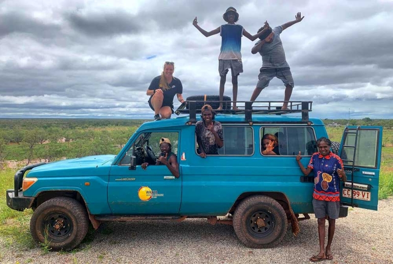 A parked blue land rover with people standing on top, hanging out of the windows and standing next to it