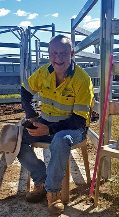 A man wearing a hi vis shirt and jeans sits in front of cattle yards smiling at the camera