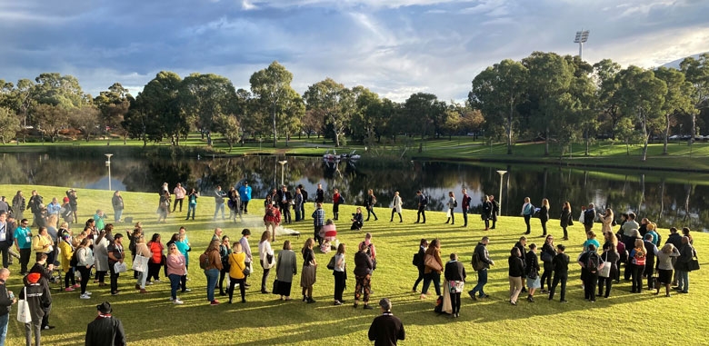 A large number of people are spread out over a lawn. In the background is a body of water and smoke rises in the middle of the group of people