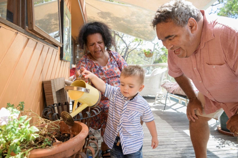 A small child waters a pot with a watering can, while 2 adults encourage them