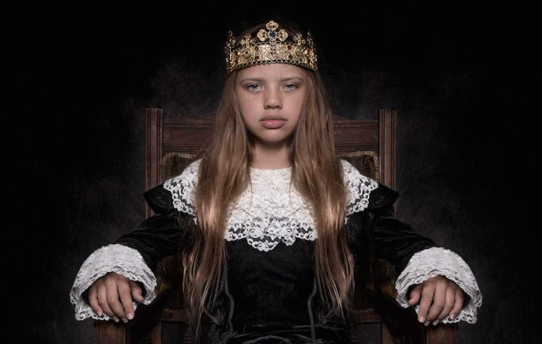 A young person in formal clothing wears a crown and sits on a chair