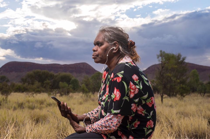 A woman sits outside with headphones in her ears connecting to the mobile phone in her hand. her eyes are closed and she looks peaceful