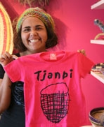 A smiling woman holds up a pink t-shirt with the word Tjanpi on it
