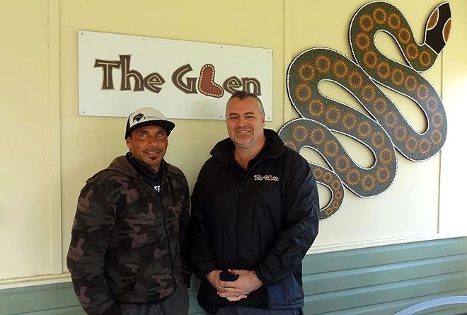 Two men in front of a wall, on which is a sign saying 'The Glen'
