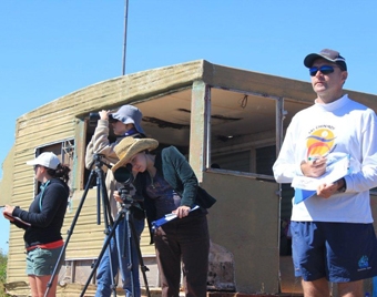 People stand near a building looking through binoculars and a camera on a tripod