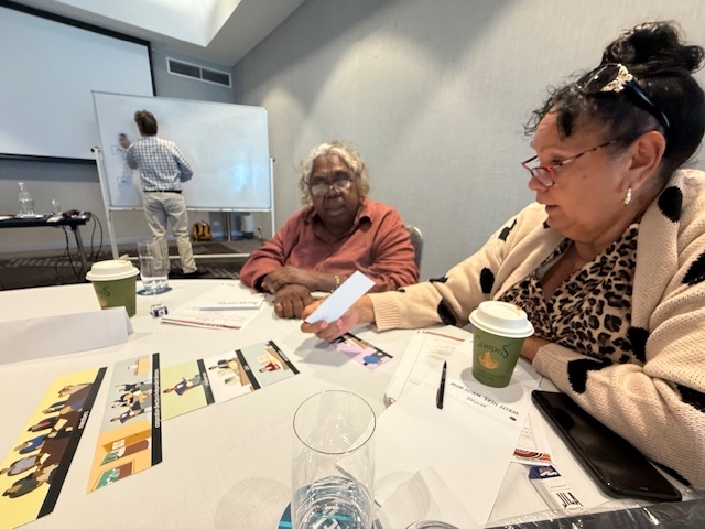 Two women sit at a table. In the background, a man is writing on a whiteboard