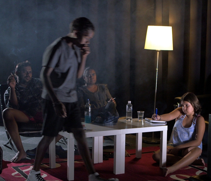 A boy walking in the foreground. In the background several adults sit on a couch and at a low table 