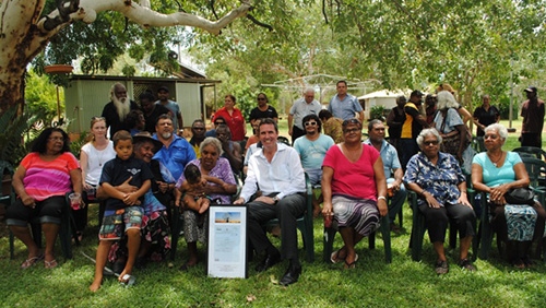 A large group of people sit and stand in an outdoor area. Two people in the front row hold a large certificate