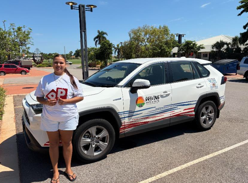 A young woman stands in front of a small white car. She is holding up 2 red P plates