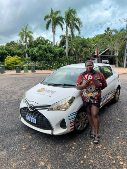 A young woman stands in front of a small white car, She is holding up red P plates