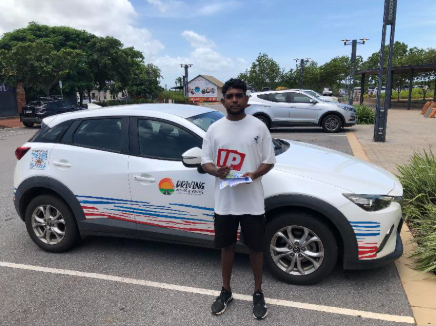 A young man stands in front of a car. He is holding up a red P plate
