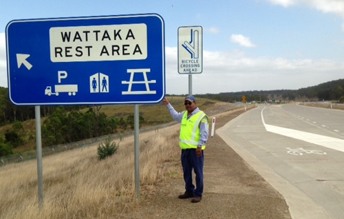 A man in a fluoro vest poses next to a road sign saying 'Whattaka rest area'