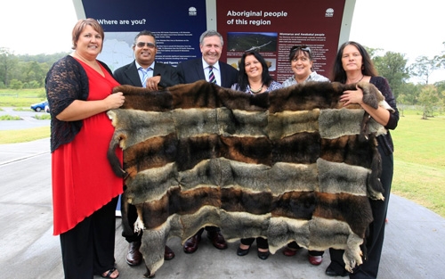 A group of people old up a large piece of fabric made of multiple grey and brown furs arranged in a striped pattern