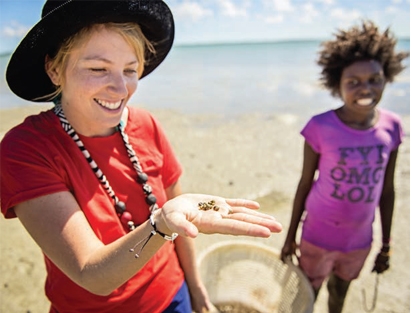 A woman on a beach holds something in her palm while the person in the background watches her smiling