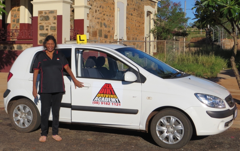 A woman stands in front of a small car with Red Dirt Driving Academy on the driver's door