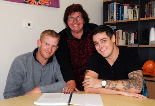 Two men sit at a table with a woman standing behind them