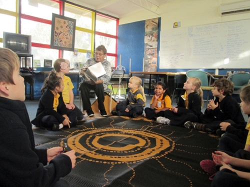 Children sit around a mat on the floor of a classroom. An adult is sitting on a chair showing them a book