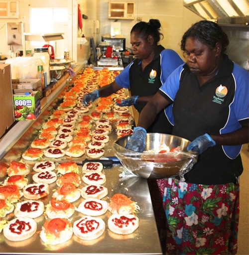 Two women add slices of tomato to rolls laid out on a large stainless steel bench
