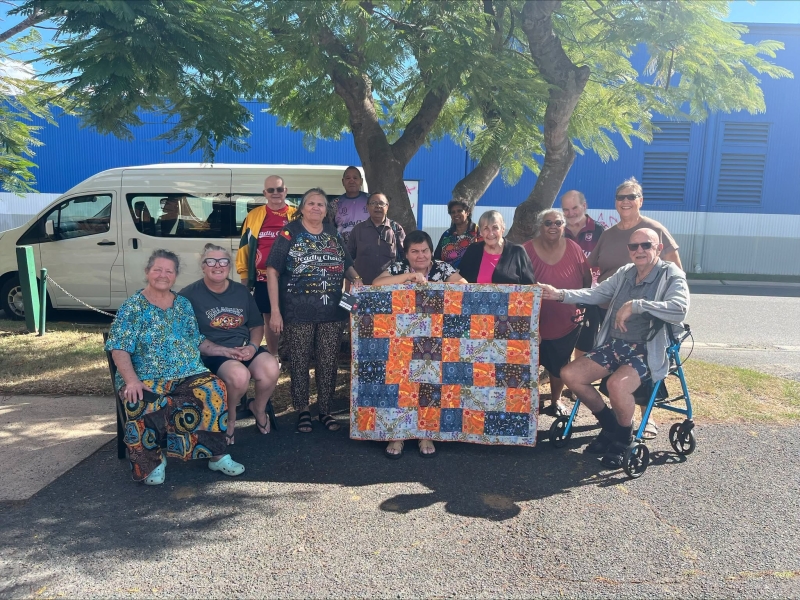 A group of people sit under a tree holding up a quilt made up of colourful squares